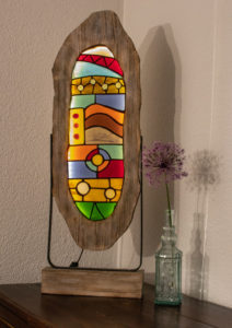 Glaslampe aus geschnittenem Buntglas in Form einer umgedrehten, fressenden Fratze. Mit einem Flachstahl auf einem Holzsockel montiert.
