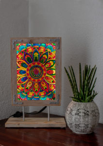 Glaslampe mit einem leuchtenden bunten auf Glas gemalten Mandala. Umrahmt von einem im Vintage-Stil gehaltenen Rahmen, der mit zwei filigranen Stahlrohren auf einen Sockel montiert ist.
