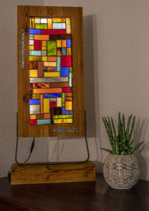 Lampe mit bunt zugeschnittenen Rechtecken aus Glas. Rahmen mit leicht orangenem Touch. Verbunden mit einem Flachstahl auf einem Holzsockel.