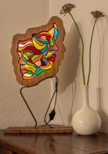 Bunte Glaslampe aus geschnittenen Buntglas, mit zwei ineinander verschmolzenen Gesichtern. Mit einem Flachstahl auf einem hölzernen Fuß montiert.